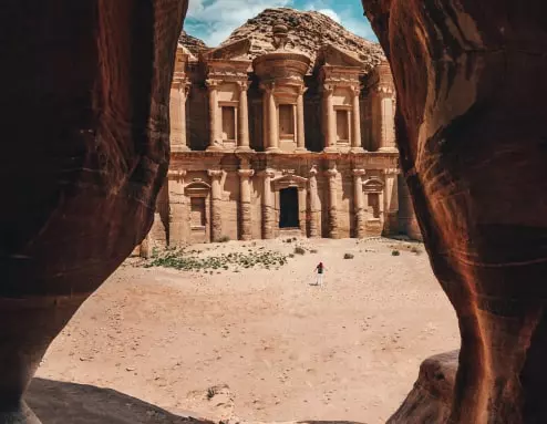 Zdjęcie przedstawia duży starożytny budynek w kolorze piasku. Można to zobaczyć po wejściu do jaskini. Pośrodku widoku znajduje się osoba.