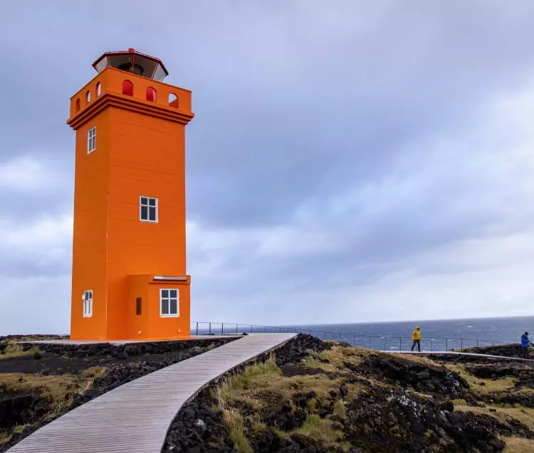Ein Foto eines quadratischen orangefarbenen Leuchtturms. Es gibt auch einen Holzweg, der zum Leuchtturm führt, auf dem sich Touristen befinden