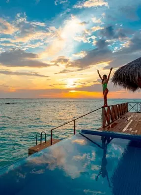 Une photo d'une jeune fille debout sur une balustrade et regardant la mer. Au premier plan il y a une piscine avec réflexion d'elle.