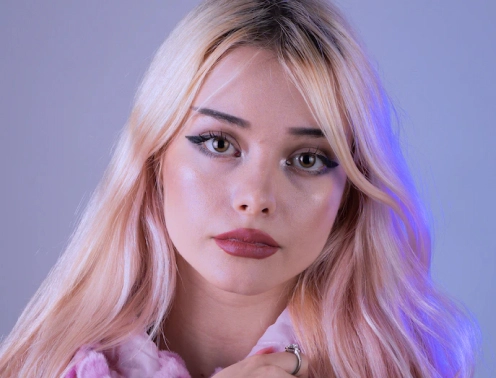 Una mujer rubia con piel pálida y lápiz labial rojo posando frente a un fondo violeta