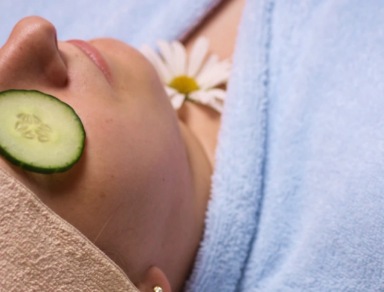 woman in a blue bathrobe laying on her back with a daisy and a cucumber slice on her face