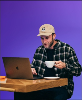 a guy in a plaid shirt and cap sits at a table in front of a laptop drinking tea