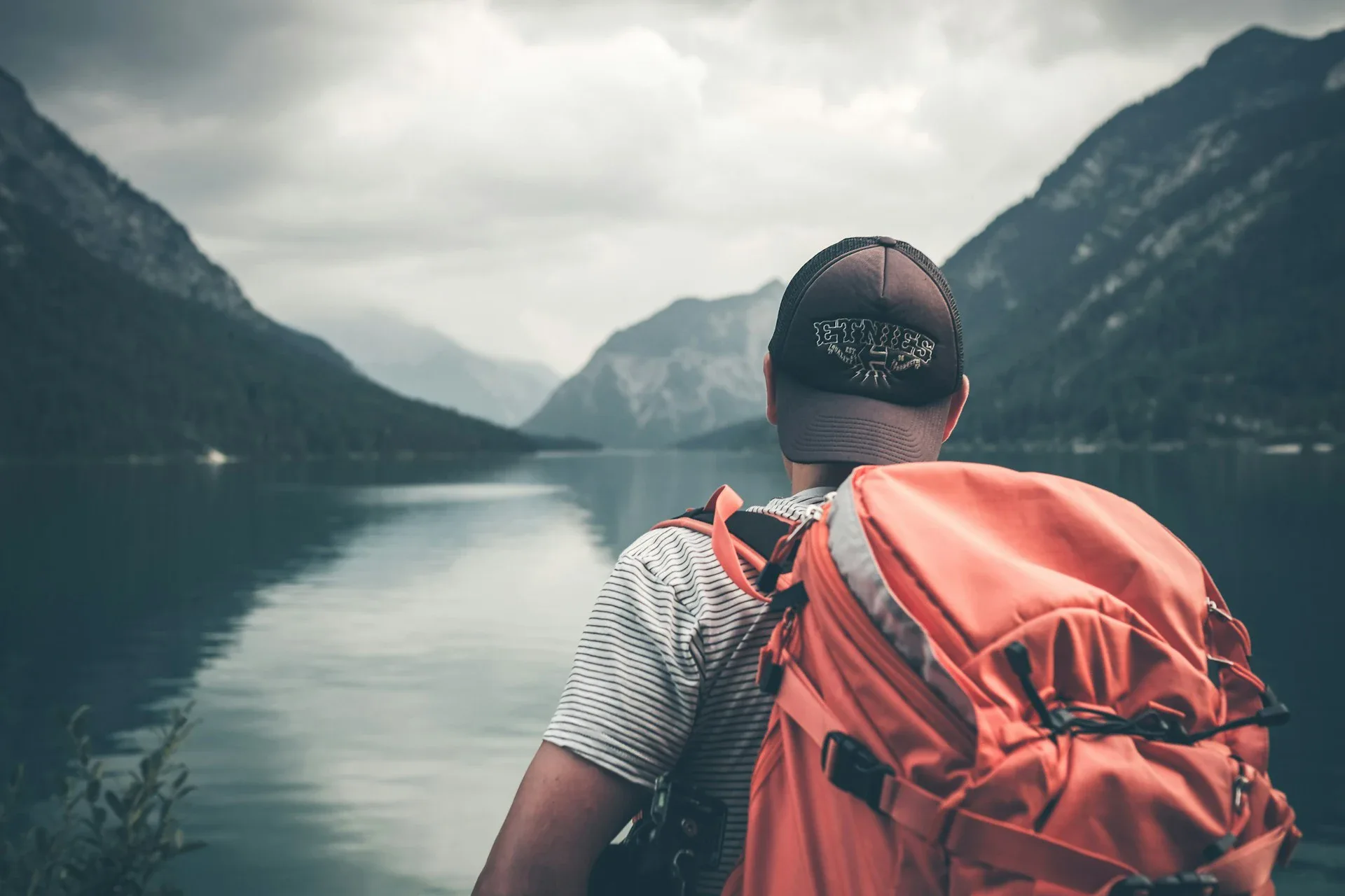 Vaikinas stovi nugara į kamerą ežero ir kalnų fone.