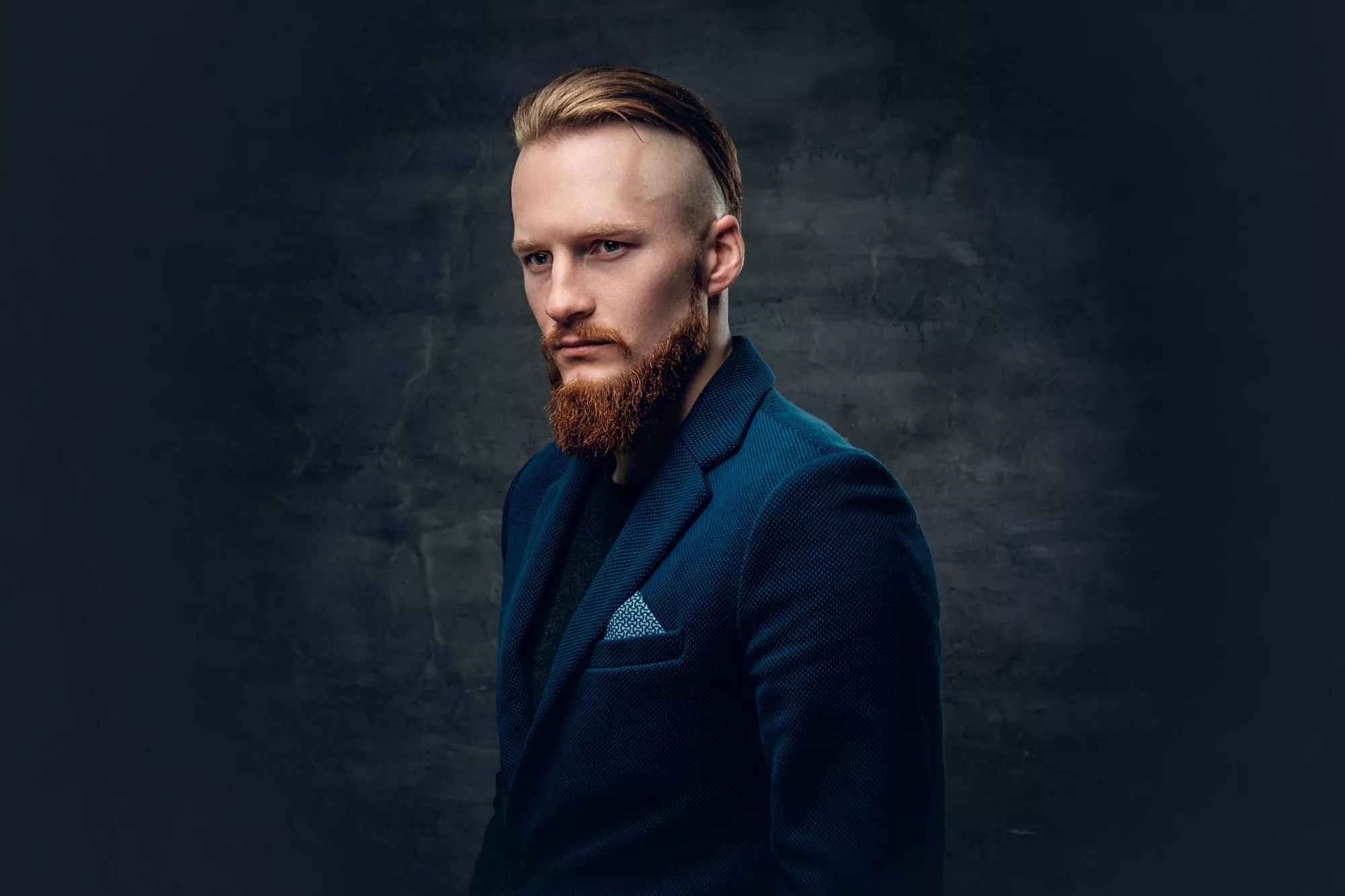 A guy with blond hair combed back and a red beard, in a dark suit with a scarf in his pocket