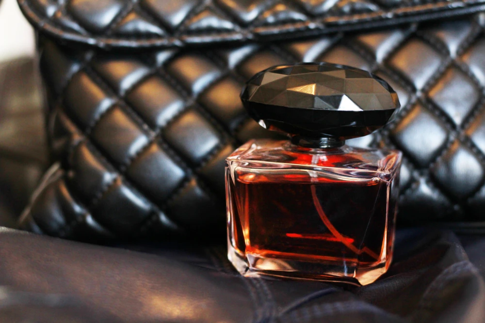 a transparent bottle of perfume stands next to a black bag with diamond stitching