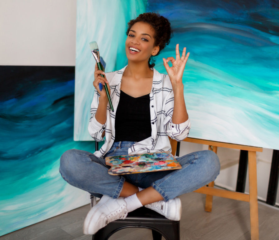 girl artist sits on a chair in the lotus position holding brushes and paint in her hands, against the background of canvas