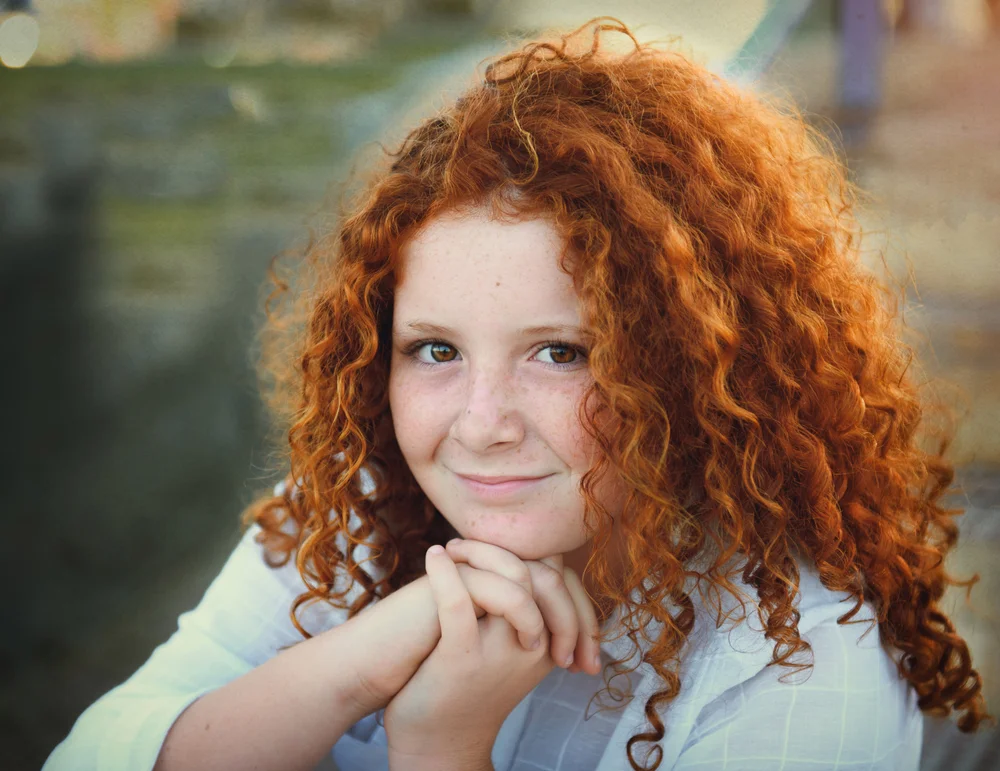 A red-haired girl with long and curly hair folded her hands in front of her face