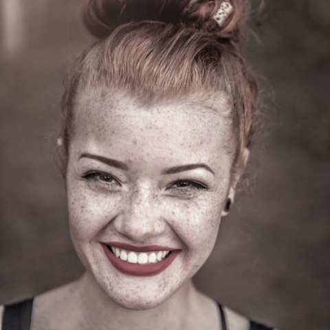 Рыжеволосая девушка с собранными волосами и коноплями на лице широко улыбается.