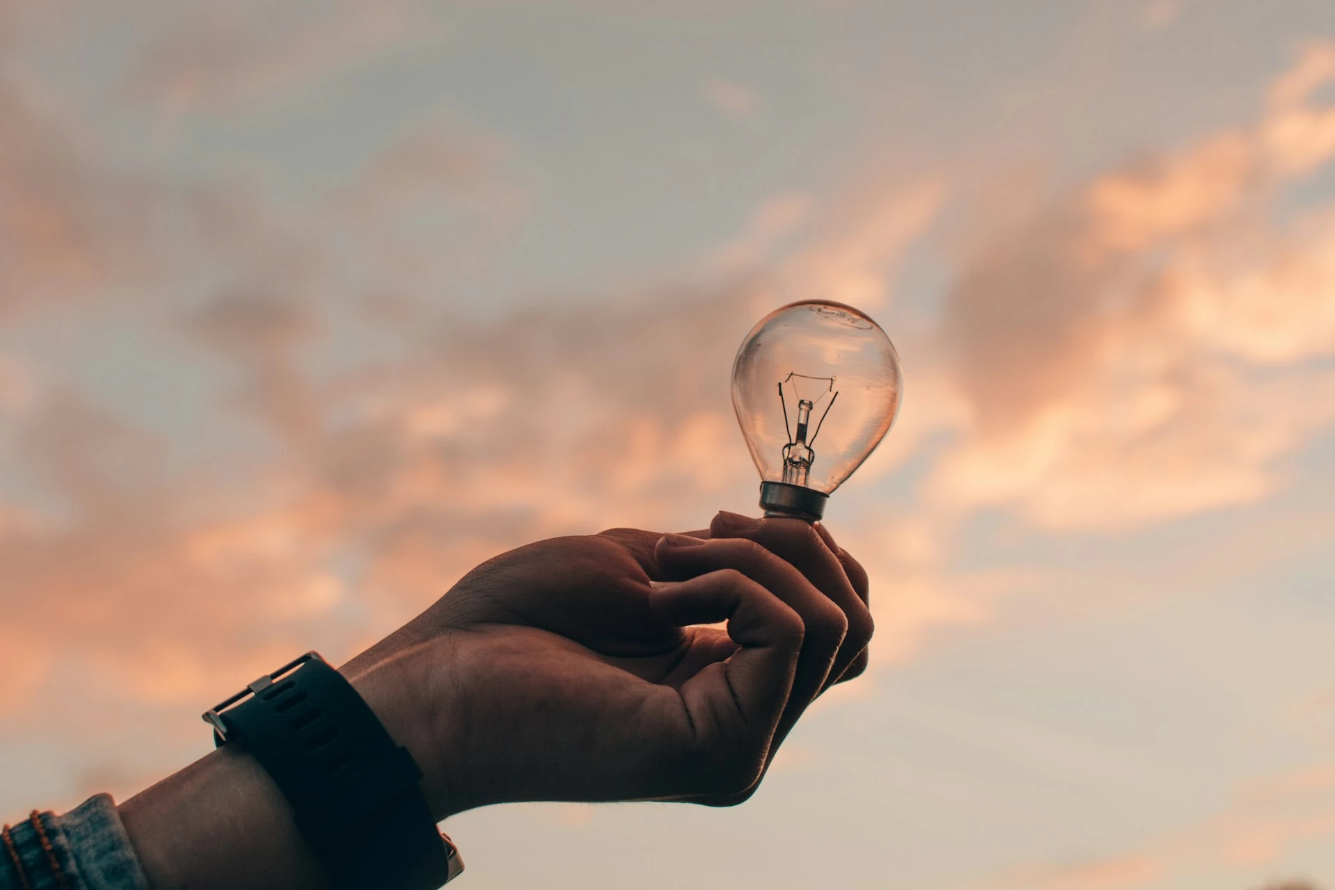 hand holding a light bulb against a background of pink and blue sky