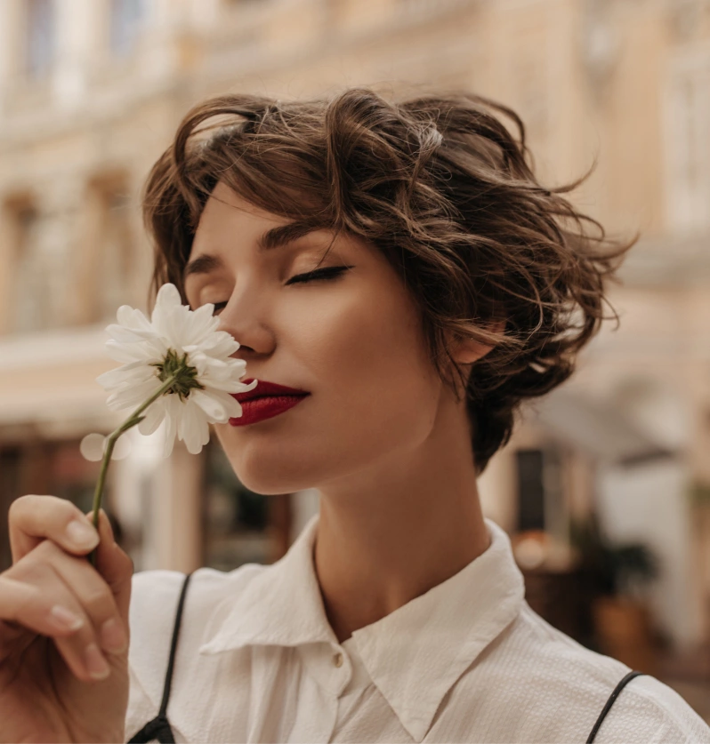 brown-haired girl with wavy hair and a short haircut smells a flower with her eyes closed